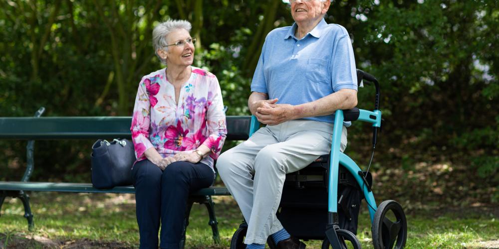 man en vrouw zitten naast elkaar zij zit op een bakje en hij op een rollator
